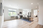 Located at 102 W Main St in Smithtown, NY, this thoughtfully designed single-floor space offers a clean, contemporary aesthetic defined by light hardwood flooring, crisp white walls, and recessed lighting throughout. The open kitchen is well-appointed with white shaker-style cabinetry extending to the ceiling, stainless steel appliances including a refrigerator, dishwasher, microwave, and range, complemented by white countertops and a undermount sink. Multiple windows flood the kitchen and adjoining dining area with natural light, creating a bright and welcoming atmosphere. The dining area accommodates a full-sized table with seating, anchored by a bold blue geometric area rug. A comfortable bedroom features neutral tones, recessed lighting, and a window with outdoor views, maintaining the space's serene character. The living area offers a minimalist setup with light wood flooring and clean sightlines. A dedicated lounge or office area includes large windows overlooking the street and vibrant blue accent seating. Two bathrooms provide distinct personalities: one features a striking blue accent wall, white vanity with stone countertop, and a backlit mirror, while the second showcases classic white subway tile, a soaking tub, walk-in shower with matte black fixtures, and a round backlit mirror — both delivering polished, modern finishes.