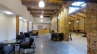 This single-floor space masterfully combines raw industrial character with thoughtful design, creating an inviting and versatile environment. Soaring ceilings showcase beautifully preserved reclaimed wood beams and exposed structural elements, lending an authentic loft aesthetic throughout. Rugged exposed brick columns and walls in warm amber tones serve as dramatic architectural focal points, contrasting elegantly with the smooth, polished concrete floors that reflect the abundant overhead lighting.

Track lighting runs along the ceiling, complementing large pendant lights that cast a warm, focused glow across the open floor plan. Natural light filters in through upper-level windows, further brightening the expansive interior. The layout is thoughtfully arranged with distinct functional zones: a lounge area features sleek black low-profile chairs, while dark wood communal tables with matching chairs provide space for collaborative gathering. A bar counter with vibrant yellow stools adds a lively pop of color, anchored by warm wooden cabinetry and decorative accents. Artwork adorns the walls, adding a curated touch to the otherwise industrial palette. The open plan and generous ceiling height create a sense of spaciousness, making this a highly functional and visually compelling space suited for a variety of uses.