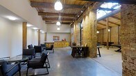 This single-floor space masterfully combines raw industrial character with thoughtful design, creating an inviting and versatile environment. Soaring ceilings showcase beautifully preserved reclaimed wood beams and exposed structural elements, lending an authentic loft aesthetic throughout. Rugged exposed brick columns and walls in warm amber tones serve as dramatic architectural focal points, contrasting elegantly with the smooth, polished concrete floors that reflect the abundant overhead lighting.

Track lighting runs along the ceiling, complementing large pendant lights that cast a warm, focused glow across the open floor plan. Natural light filters in through upper-level windows, further brightening the expansive interior. The layout is thoughtfully arranged with distinct functional zones: a lounge area features sleek black low-profile chairs, while dark wood communal tables with matching chairs provide space for collaborative gathering. A bar counter with vibrant yellow stools adds a lively pop of color, anchored by warm wooden cabinetry and decorative accents. Artwork adorns the walls, adding a curated touch to the otherwise industrial palette. The open plan and generous ceiling height create a sense of spaciousness, making this a highly functional and visually compelling space suited for a variety of uses.