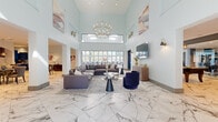 The 2nd Phase Clubhouse in Round Rock, TX, is a expansive community facility thoughtfully designed to offer a premium social and recreational experience. The centerpiece of the space is a grand lounge and living area boasting soaring double-height ceilings, marble-look tile flooring, and an abundance of natural light streaming through large floor-to-ceiling windows. A stunning tiered chandelier anchors the room, which is furnished with plush sectional sofas, accent chairs, and curated artwork, creating an inviting and sophisticated atmosphere. Adjacent to the lounge, a well-appointed kitchen features bold navy-blue cabinetry with gold hardware, white quartz countertops, herringbone tile backsplash, and stainless steel appliances, flowing seamlessly into a dining area. The fitness center is bright and modern, lined with floor-to-ceiling windows overlooking the outdoors and outfitted with cardio equipment and free weights. A game room with a billiard table, bold blue accent wall, and mirrored wall adjacent to additional workout equipment adds a dynamic recreational element. A private conference or office room provides a quiet workspace. Three well-finished bathrooms feature marble-look tile and contemporary fixtures. Hallways connect each area efficiently, and pool access is visible through glass doors, extending the facility's amenities outdoors.