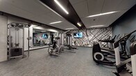 Located at 590 Park St in St. Paul, Minnesota, this modern fitness center is a well-equipped, single-floor facility designed to support a comprehensive workout experience. The space is divided into distinct functional zones, each thoughtfully arranged to accommodate a variety of exercise disciplines.

The strength training area is anchored by a selection of Life Fitness selectorized machines, including chest press and leg press units, complemented by a full dumbbell rack offering a wide range of free weights. Large wall-to-wall mirrors line one side of the room, enhancing the sense of space and allowing users to monitor their form throughout their workouts. Mounted flat-screen TVs are positioned for easy viewing across the floor.

A dedicated cardio zone features multiple elliptical trainers, upright bikes, and a rowing machine, all set against a deep steel-blue accent wall that adds a cool, focused atmosphere to the space. A water station with a trash receptacle is conveniently located nearby.

Throughout the facility, linear LED lighting runs across the suspended ceiling panels, providing bright, even illumination. The standout design element is a striking black-and-white street map mural that wraps across a prominent wall, serving as both a bold artistic statement and a nod to the surrounding St. Paul cityscape. Durable, multi-tone carpet tile flooring completes the polished, professional aesthetic of this well-appointed fitness facility.