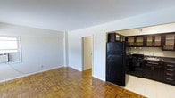 This single-floor unit showcases a practical and straightforward layout designed to maximize usable space. The main living area is characterized by warm-toned parquet hardwood flooring arranged in a classic herringbone-style pattern, adding a timeless aesthetic to the room. Walls are finished in a clean, neutral white, creating a bright and open atmosphere that complements the natural tones of the flooring.

An open archway connects the living area to the kitchen, providing a sense of flow between the two spaces. The kitchen is equipped with black appliances, including a full-size refrigerator and a gas range, and features dark upper cabinetry offering ample storage. The kitchen floor transitions to light-toned tile, providing a clear visual distinction between the two areas.

A doorway visible from the main living area leads to an additional room, suggesting further functional living space within the unit. The smooth white ceilings and simple baseboard trim throughout contribute to a clean, understated interior aesthetic. Overall, the space offers a no-frills, functional environment well-suited for everyday living.