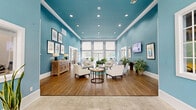 Located at 1767 Hermitage Blvd in Tallahassee, Florida, this single-floor community clubhouse serves as a vibrant gathering hub designed with both comfort and functionality in mind. The space is immediately distinguished by its soaring ceilings adorned with crisp white crown molding that contrasts beautifully against bold teal walls, creating an energetic yet welcoming ambiance throughout.

The main lounge area features warm hardwood-style flooring and is furnished with a curated arrangement of neutral-toned armchairs and sofas centered around a round coffee table, ideal for relaxed socializing. Recessed lighting supplements the generous natural light that pours in through large, multi-pane windows, offering views of the surrounding greenery. A wall-mounted flat-screen television and carefully placed potted plants add to the livability of the space.

A glass-paneled office or leasing area is accessible directly from the main lounge, providing a transparent and professional workspace visible from the common area. An adjacent conference or meeting room, also enclosed in glass, offers a more structured setting for group activities or business needs. Additional high-top bar seating near the windows provides an alternative workspace. A fitness center with cardio equipment is accessible through a dedicated doorway, rounding out the amenities. A welcoming entrance mat greets visitors upon arrival through the exterior glass doors.