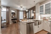 This single-floor residence is designed with an open-concept layout that seamlessly connects the kitchen and living area. The kitchen is a standout feature, equipped with stainless steel appliances including a gas range, microwave, refrigerator, and dishwasher. White shaker-style cabinetry with frosted glass uppers pairs with striking granite countertops, while a large central island with a farmhouse sink and pendant lighting serves as both a functional workspace and a social hub. A decorative arabesque tile backsplash adds a refined touch, and warm hardwood flooring flows throughout the main living areas.

The living space transitions naturally from the kitchen and is brightened by natural light through slatted blinds and patterned curtains. Both bedrooms feature plush carpet, ceiling fans, and generous windows. The primary bedroom is distinguished by a bold teal accent wall adorned with circular wall art, lending a sophisticated and artistic character to the space.

The bathroom offers a long, functional layout with dual under-mount sinks set into dark granite countertops, white cabinetry with brushed nickel hardware, and a tub-shower combination. Recessed and sconce lighting provide ample illumination throughout this well-appointed residence.