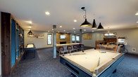 This single-floor recreational space is thoughtfully designed to serve as a premier entertainment hub. At the heart of the room sits a full-size pool table, illuminated by a trio of sleek black pendant lights that draw the eye and anchor the space with modern industrial flair. Recessed ceiling lighting throughout ensures the entire area is well-lit and inviting.

A central bar counter with stylish wooden barstools provides a natural gathering spot, complemented by a wall-mounted flat-screen TV set against a warm wood-paneled accent wall — creating a cozy focal point ideal for watching games or socializing. Adjacent to the bar, a dining area with additional seating extends the space's functionality.

To one side, a classic arcade game adds a playful, nostalgic touch, making this space as versatile as it is entertaining. The flooring features a durable, textured dark carpet that ties the room together while offering comfort underfoot. Neutral gray walls and structural columns give the space a clean, contemporary aesthetic. Windows along the perimeter allow natural light to filter in, brightening the atmosphere. Overall, this space is purpose-built for recreation and community, combining comfort, style, and a wide range of leisure amenities.