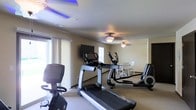 This single-floor fitness center offers a dedicated space for cardiovascular exercise and relaxation. The room is outfitted with a selection of quality cardio equipment, including a treadmill, an elliptical trainer, and a stationary bike, providing a comprehensive range of workout options for users of varying fitness levels. A wall-mounted flat-screen television is positioned for easy viewing during workouts, adding an entertainment element to exercise sessions. The space is illuminated by ceiling fans with integrated light fixtures, which contribute to comfortable airflow and consistent lighting throughout. A distinctive blue ambient light accent near the ceiling adds a modern, energizing touch to the room's atmosphere. Natural light filters in through windows dressed with sheer white curtains, creating a bright and welcoming environment. The flooring is finished with neutral-toned carpet, offering a soft underfoot surface suitable for fitness use. A small table and chair set in the background provides a convenient area to rest or review workout plans. The overall ambiance is clean, functional, and inviting — well-suited for daily fitness routines.
