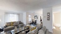 This single-floor residence showcases an open-concept design that seamlessly integrates the living, dining, and kitchen areas into one cohesive and functional space. The living area is anchored by a comfortable seating arrangement featuring grey upholstered sofas accented with patterned and mustard-yellow throw pillows, centered around a sleek glass coffee table. Natural light fills the space through multiple windows dressed with sheer white curtains, creating an airy and inviting atmosphere throughout.

The dining area transitions naturally from the living space, furnished with a round dark table surrounded by industrial-style metal chairs, set atop a patterned area rug. A modern pendant light hangs overhead, adding a contemporary focal point to the dining zone. Beyond the dining area, a glimpse into the kitchen reveals dark cabinetry, offering a contrast to the neutral white walls found throughout the home.

The textured popcorn ceiling and warm wood-tone flooring lend a classic character to the space, while the open layout maximizes the sense of spaciousness. The overall ambiance is clean, modern, and thoughtfully styled with layered décor and cohesive neutral tones accented by bold geometric patterns.