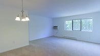 This single-floor residence features an open and airy living area finished with wall-to-wall neutral carpeting that adds warmth to the space. The room is bathed in natural light through a large window that frames a pleasant view of lush, mature trees outside, creating a connection to the outdoors. White walls throughout provide a clean, bright backdrop that enhances the sense of openness. A classic multi-bulb chandelier with a brass finish anchors one end of the room, adding a traditional touch to the overhead lighting. Climate control is provided by a wall-mounted air conditioning unit, complemented by a baseboard heating system that runs along the exterior wall, ensuring year-round comfort. The layout offers generous open floor space, making the area well-suited for a variety of furniture arrangements. The overall ambiance is bright and neutral, presenting a functional and move-in-ready environment with practical amenities integrated seamlessly into the design.