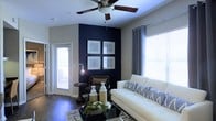 This single-floor space showcases a thoughtfully designed interior that balances modern aesthetics with comfortable living. The living area serves as the centerpiece, featuring a striking navy blue accent wall that creates a bold focal point and adds depth to the room. Four framed abstract artworks are displayed symmetrically on the accent wall, reinforcing the space's contemporary character.

The room is furnished with a clean-lined white sofa accented with patterned throw pillows, complemented by a sleek glass coffee table and a dark leather armchair. Dark hardwood flooring runs throughout, providing a rich contrast to the light-toned walls and bright furnishings. A ceiling fan with a brushed metal finish is centrally mounted, adding both function and a refined visual detail overhead.

Natural light pours in generously through large windows dressed with horizontal blinds and grommet-style curtain panels, creating a bright and airy atmosphere. A glass-paneled door provides additional light and a view to the exterior. Subtle ambient lighting from a modern Edison-style floor lamp adds warmth to the seating area. Overall, the space exudes a polished, contemporary ambiance with a cohesive color palette of navy, white, and charcoal.