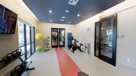 This fitness facility is designed to support a well-rounded workout experience within a clean, modern environment. The space is defined by its distinctive navy blue ceiling, which contrasts sharply with the light-colored walls and creates a focused, energized atmosphere. Recessed lighting is evenly distributed throughout the ceiling, ensuring bright, consistent illumination across the entire area.

The flooring is a neutral tone accented by a bold red runner strip down the center, visually dividing the space and adding a dynamic design element. A set of glass-paneled double doors leads to an adjoining room visible in the background, which appears to house additional strength and resistance training equipment, effectively expanding the overall fitness offerings.

In the main area, stationary cycling bikes are positioned centrally, making them a focal point of cardio training. To one side, a colorful collection of stability and medicine balls is neatly organized on a storage rack, offering versatile options for functional fitness. Large windows along the perimeter allow natural light to filter in, contributing to an open and inviting atmosphere. The layout is thoughtfully arranged to allow freedom of movement throughout.