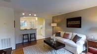 This single-floor space offers a thoughtfully designed open-concept layout that blends comfort and functionality. The living area serves as the heart of the home, featuring warm hardwood flooring, neutral gray walls, and a large sectional sofa complemented by accent pillows and a bold geometric area rug. A sleek dark-finished coffee table anchors the seating arrangement, while a tall floor lamp provides warm ambient lighting. A striking piece of wall art adds a pop of color and visual interest to the space.

A defining architectural feature is the elegant arched entryway, fitted with track lighting, that leads seamlessly into the kitchen and breakfast bar area. The kitchen is bright and well-lit, benefiting from natural light streaming through a window, and includes white cabinetry and ample counter space. The breakfast bar, lined with two classic black bar stools, creates an inviting space for casual dining or entertaining.

The overall ambiance is warm and welcoming, with a cohesive neutral color palette punctuated by thoughtful design details. The open flow between the living and kitchen areas makes the space feel connected and spacious, ideal for both everyday living and hosting guests.