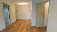 This single-floor space offers a clean, functional layout characterized by warm, richly toned hardwood flooring that extends throughout the main living areas, lending a sense of continuity and warmth. The neutral, light-toned walls create a bright and open atmosphere, while the textured ceiling adds a subtle architectural detail overhead. Overhead track lighting in the main living area provides ample illumination, making the space feel well-lit and inviting even without relying solely on natural light. The layout features multiple interior doorways, offering clear delineation between the different areas of the space — including what appears to be a utility or entry passage and additional adjoining rooms. A partial wall with an open pass-through connects the living area to what appears to be a kitchen or adjacent space, hinting at an open-concept flow between functional zones. The overall ambiance is clean and move-in ready, with a straightforward design that provides a versatile canvas for a variety of uses. The combination of hardwood floors, neutral palette, and practical room transitions gives this space a welcoming, understated character suited to everyday living.