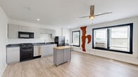 Located at 511 Maple Blvd in Kansas City, MO, this studio unit offers an efficient, thoughtfully designed single-floor layout combining a kitchen and living area with one full bathroom. The open-plan living and kitchen space is anchored by light wood-look flooring that runs throughout, creating a clean and cohesive aesthetic. The kitchen is outfitted with stainless steel appliances, including a refrigerator, electric range, over-the-range microwave, and dishwasher, complemented by dark countertops and modern gray cabinetry. A movable kitchen island with a butcher block top provides added prep and dining flexibility. The living area is bright and open, illuminated by natural light streaming through two dark-framed windows fitted with blinds. A ceiling fan with integrated lighting serves as a central fixture overhead. A distinctive feature of the living space is a section of exposed original brick on the interior wall, adding character and a nod to the building's history. The bathroom is clean and functional, featuring a walk-in shower stall, a wall-mounted vanity with a wood-toned cabinet, and matching wood-look flooring. White walls throughout keep the space feeling bright and open, while recessed lighting adds a modern touch to the kitchen area.