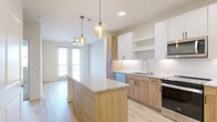 This well-designed single-floor residence offers a bright, contemporary living experience through a thoughtful open-concept layout. The kitchen serves as the centerpiece of the space, showcasing warm natural wood cabinetry paired with crisp white upper cabinets, granite countertops, and a full suite of stainless steel appliances including a refrigerator, range, microwave, and dishwasher. A central kitchen island with a matching granite surface provides additional workspace and casual dining potential, illuminated by stylish amber glass pendant lights. A white subway tile backsplash and under-cabinet lighting add refined detail throughout.

Light wood-tone flooring runs through the main living and kitchen areas, contributing to the space's airy, modern feel. The open layout flows seamlessly toward a set of glass French doors that open to a private balcony, allowing natural light to fill the interior. A ceiling fan in the living area provides year-round comfort.

The bedrooms feature soft neutral carpeting and are well-lit by large windows with views to the surrounding area. The bathroom is finished with light wood-tone flooring, a white vanity with mirror, and a tub-shower combination with tile surround. Completing the unit's practical offerings is a conveniently located in-unit stacked washer and dryer, providing day-to-day functionality within the residence.