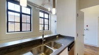 This 1-bedroom, 1-bathroom floor plan showcases a compelling blend of industrial and contemporary design elements. The kitchen is a standout feature, equipped with a double-basin stainless steel sink paired with a sleek, professional-style faucet. Dark stone countertops provide generous prep space and contrast beautifully against the lighter cabinetry below. Black-framed windows allow abundant natural light to fill the space while framing views of the surrounding brick exterior architecture, reinforcing the unit's urban character. Overhead, exposed metallic ductwork runs along the ceiling, a nod to the building's industrial heritage, complemented by cylindrical pendant lighting that adds focused illumination and a modern touch. The white walls and clean lines balance the rawness of the industrial elements, creating a space that feels both functional and stylish. The single-floor layout ensures a straightforward, accessible living arrangement. Overall, the unit delivers a cohesive loft-inspired aesthetic, combining durable, low-maintenance materials with thoughtful design details suited for contemporary urban living.