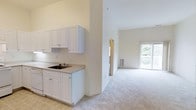 This 2-bedroom, 2-bathroom single-floor residence offers an open, airy layout defined by soaring vaulted ceilings that lend a sense of spaciousness throughout. The kitchen features white cabinetry with classic raised-panel doors, a double-basin sink, and light countertops, with tile flooring underfoot. The space transitions seamlessly into the main living area, which is finished with neutral wall-to-wall carpeting that adds warmth and comfort. The living room benefits from abundant natural light streaming through a large window that frames an outdoor view of a covered balcony or patio, enhancing the connection between the interior and exterior. The consistent light and neutral color palette — cream walls, white trim, and soft flooring — creates a clean, cohesive ambiance throughout the home. The single-floor design ensures easy accessibility and a straightforward flow between rooms. Overall, the space offers a comfortable and functional environment well-suited for everyday living.