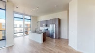 This contemporary single-floor residence showcases a thoughtfully designed open-concept layout that seamlessly connects the kitchen and living areas. The kitchen is a standout feature, equipped with stainless steel appliances including a refrigerator, electric range, and built-in microwave, complemented by sleek dark wood cabinetry, a white subway tile backsplash, and a central island with a dark stone countertop and undermount sink. Light-toned hardwood flooring flows throughout the main living areas, enhancing the bright, airy atmosphere created by an impressive wall of floor-to-ceiling windows. These expansive black-framed windows flood the space with natural light and frame sweeping views of the surrounding cityscape and distant mountains. The bedrooms offer plush carpeting and generous wall space, with at least one room also benefiting from large floor-to-ceiling windows overlooking the urban landscape. The bathrooms are finished with large dark tile flooring, wood-toned vanities with stone countertops, and decorative sconce lighting. One bathroom features a walk-in glass-enclosed shower adorned with patterned tile, while another includes a soaking tub combo. The overall design is clean, modern, and cohesive throughout.