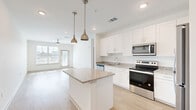 This thoughtfully designed single-floor residence offers a clean, contemporary aesthetic highlighted by a bright, neutral color palette and light wood-style flooring that flows seamlessly through the main living areas. The open-concept kitchen is a standout feature, anchored by a central island with granite countertops and illuminated by a pair of stylish pendant lights. White shaker cabinetry lines the walls, complemented by a full suite of stainless steel appliances including a refrigerator, electric range, microwave, and dishwasher. The kitchen opens directly into the living area, which benefits from abundant natural light streaming through large windows and a glass door leading to a private outdoor access point. A ceiling fan adds comfort to this airy, open space. The bedrooms are carpeted for warmth and comfort, each featuring ceiling fans and windows that invite natural light. The bathrooms maintain the same refined design language found throughout the home, with granite-topped white vanities and wood-style tile flooring. One bathroom includes a tub-shower combination with clean white tile surround. The overall ambiance of the space is modern, light-filled, and move-in ready, with consistent finishes that create a cohesive and welcoming environment.