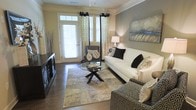 This single-floor living space showcases a thoughtfully designed interior that balances comfort and sophistication. The living area serves as the focal point, featuring rich hardwood flooring that anchors the room with warmth and durability. Crown molding adds a refined architectural detail along the ceiling line, enhancing the overall sense of polish throughout the space.

Natural light flows generously into the room through a glass-paneled entry door fitted with white plantation shutters, complemented by tall windows dressed in decorative patterned drapes with grommet tops in a black and neutral palette. The combination creates an inviting, well-lit atmosphere while maintaining privacy options.

The living room is furnished with a clean-lined white sofa accented by black and neutral throw pillows, paired with patterned accent chairs and a striking round glass coffee table with a sculptural base. A large area rug in muted earth tones defines the seating arrangement, while a dark media console anchors one wall. A bold abstract canvas in blue and grey tones adds a standout artistic element, contributing to the space's contemporary yet welcoming ambiance.