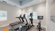 Located at 5901 Myrtle Dr in Wildwood, Florida, this fitness center offers a well-equipped, single-room facility designed to support a comprehensive workout experience. The space features a distinctive tray ceiling with recessed lighting that enhances the sense of openness, while white walls and neutral tones create a clean, modern aesthetic throughout.

The flooring is a patterned commercial carpet with bold gray, teal, and orange stripe accents, adding a dynamic visual element to the space. Natural light filters in through multiple white-shuttered windows along the walls, contributing to an inviting and energizing atmosphere.

The cardio zone is equipped with treadmills, an elliptical trainer, and a stationary bike with an integrated screen, catering to a range of cardiovascular training preferences. The strength training area includes a multi-functional cable machine, a leg press, a dumbbell rack with a full set of free weights, adjustable benches, and additional resistance equipment. Floor-to-ceiling mirrors along one wall visually expand the space and allow users to monitor their form during workouts.

A ceiling fan provides additional air circulation, and the facility includes proper exit signage and ventilation. Signage identifying the space as a Fitness Center is also present near the entrance, which connects to a hallway beyond.