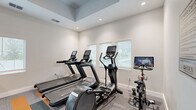 Located at 5901 Myrtle Dr in Wildwood, Florida, this fitness center offers a well-equipped, single-room facility designed to support a comprehensive workout experience. The space features a distinctive tray ceiling with recessed lighting that enhances the sense of openness, while white walls and neutral tones create a clean, modern aesthetic throughout.

The flooring is a patterned commercial carpet with bold gray, teal, and orange stripe accents, adding a dynamic visual element to the space. Natural light filters in through multiple white-shuttered windows along the walls, contributing to an inviting and energizing atmosphere.

The cardio zone is equipped with treadmills, an elliptical trainer, and a stationary bike with an integrated screen, catering to a range of cardiovascular training preferences. The strength training area includes a multi-functional cable machine, a leg press, a dumbbell rack with a full set of free weights, adjustable benches, and additional resistance equipment. Floor-to-ceiling mirrors along one wall visually expand the space and allow users to monitor their form during workouts.

A ceiling fan provides additional air circulation, and the facility includes proper exit signage and ventilation. Signage identifying the space as a Fitness Center is also present near the entrance, which connects to a hallway beyond.