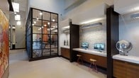 This well-appointed amenity space combines functionality and modern design across a single floor. The fitness center is prominently featured behind a striking floor-to-ceiling steel-framed glass partition, offering a transparent view into the workout area while maintaining a sense of openness. Inside, the gym is outfitted with a variety of cardio and strength training equipment, accented by warm pendant lighting and a bold architectural column that adds a distinctive visual element to the space.

Adjacent to the fitness center is a dedicated business center workstation, thoughtfully designed with a long, wood-toned built-in desk that comfortably accommodates multiple users. The workstation is framed by an eye-catching textured stone accent wall, which adds warmth and depth to the contemporary aesthetic. Under-cabinet lighting illuminates the desk area, creating a focused and productive environment. Computer monitors are built into the station, making it a ready-to-use workspace.

The overall design language is cohesive and sophisticated, blending dark wood cabinetry, neutral flooring, and clean architectural lines. The combination of the transparent gym wall and the richly textured accent surfaces creates an inviting and polished communal environment ideal for both fitness and productivity.