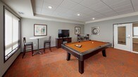 This single-floor amenity space is thoughtfully designed to serve as a social hub, combining recreation, comfort, and functionality in one cohesive environment. The space is unified by a bold, patterned carpet in striking shades of orange and gray, complemented by warm terracotta-toned accent walls that lend energy and personality throughout.

The game room is a standout feature, centered around a full-size pool table with cue storage mounted on the wall. A large flat-screen television, high-top bar tables with stools, and framed artwork complete the space, making it ideal for casual gatherings and entertainment.

Adjacent to the game room, a generously sized lounge area offers comfortable sectional and accent seating arranged to encourage conversation. Roller shades on large windows allow for controlled natural light, while glass doors provide direct access to an outdoor area, enhancing the sense of openness.

The kitchenette is sleek and well-appointed, featuring dark wood cabinetry, dark granite countertops, and a luminous textured tile backsplash. Stainless steel appliances—including a full-size refrigerator, dishwasher, and microwave—make the space fully functional for hosting events. Recessed lighting and a neutral floor tie the kitchen area together with clean, modern finishes throughout.