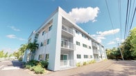 This contemporary residential building presents a striking modern aesthetic, characterized by its clean white stucco exterior and well-proportioned facade. The structure rises across multiple levels, each floor featuring private balconies outfitted with minimalist metal railings that contribute to the building's streamlined architectural character. Large windows are evenly distributed across the exterior, designed to welcome natural light into each unit while offering outward views of the surrounding environment. The building's exterior is complemented by mature tropical plantings and palms along its base, softening the crisp architectural lines and adding a lush, verdant contrast to the bright white facade. The overall design reflects a clean, modern sensibility well-suited to a warm-weather setting, with an emphasis on outdoor connectivity through the private balcony spaces. The building's layout suggests thoughtfully organized floor plans on each level, with consistent design elements carried throughout. The bright, sunny setting and tropical greenery surrounding the structure contribute to an inviting, resort-like atmosphere, making this a distinctive and well-presented residential property.
