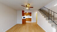 Located at 6150 SW King Blvd in Beaverton, Oregon, this two-floor residence offers a practical and well-organized layout across two equally sized floors. The main level features an open-plan living, dining, and kitchen area finished with light wood laminate flooring throughout. The kitchen is equipped with warm-toned wood cabinetry, black appliances including a refrigerator and electric range with hood, a double-basin sink, and ample counter space. A ceiling fan with integrated lighting anchors the living area, which also includes a wall-mounted HVAC unit. A large front-facing window allows natural light to fill the space, and a front entry door provides direct exterior access. A staircase with metal railing connects the main level to the upper floor. The upper level is carpeted and serves as the primary bedroom, offering generous square footage along with a dedicated wall HVAC unit and a window with an outdoor view. Two bathrooms are present in the home, each featuring warm wood vanity cabinetry, white countertops, a sink with brushed nickel fixtures, and overhead vanity lighting. One bathroom includes access to a dedicated closet with shelving. Textured ceilings are consistent throughout both floors, contributing to the home's cozy and functional character.