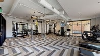 Located at 1005 W Main St in Durham, NC, this contemporary fitness center offers a thoughtfully designed, single-floor facility built to support a wide range of workout routines. The main gym area is defined by a striking herringbone-patterned carpet floor in neutral tones of gray, beige, and white, lending a polished, modern aesthetic throughout the space. Exposed white ductwork runs along the ceiling alongside ceiling fans and stylish drum pendant lights, creating an industrial-chic ambiance that feels both functional and refined. Full-length mirrors line one wall, enhancing the sense of openness.

The facility is comprehensively equipped with strength training stations including a multi-functional power rack with TRX suspension straps, free weights, and cable machines, as well as cardiovascular equipment such as treadmills, ellipticals, stair climbers, and stationary bikes. Floor-to-ceiling windows along the perimeter flood the space with natural light and frame views of Durham's historic architecture, including neighboring brick buildings that reflect the city's character.

A separate studio room features light wood-plank flooring, a ballet barre, cubbies for personal items, and space for group or individual classes. Wall-mounted televisions are positioned throughout for entertainment during workouts. The overall design balances high-end finishes with a practical, welcoming environment suited to serious fitness pursuits.