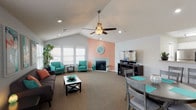 This single-floor space makes a strong first impression with its vaulted ceilings that lend an airy, expansive feel to the main living area. A ceiling fan is centrally mounted at the peak of the cathedral ceiling, providing both function and visual interest. The living room is anchored by a fireplace set against a warm peach accent wall, above which a decorative round mirror adds a stylish focal point. Large windows along one wall allow natural light to fill the room, enhancing the open atmosphere. The space is finished with plush wall-to-wall carpeting in a neutral tone, providing a comfortable and cohesive foundation throughout. The living area is furnished with a dark sofa, a classic X-frame coffee table, and a pair of teal accent chairs that inject a lively pop of color. A flat-screen TV and media console are positioned opposite the seating area, completing the entertainment setup. Seamlessly adjoining the living room is a dedicated dining area, featuring a compact dining table with gray chairs accented by teal placemats, maintaining the consistent color theme. Recessed lighting and a textured ceiling further complement the overall design. The result is a cohesive, well-lit space that balances comfort and style.