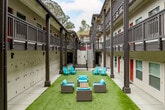 Located at 630 Water St in Santa Cruz, California, this residential complex is centered around a vibrant, elongated courtyard that serves as the heart of the community. The courtyard is lined with lush artificial turf, creating a clean, low-maintenance green space that runs the length of the building. Bright teal Adirondack chairs and wicker lounge seating with turquoise cushions are arranged throughout the courtyard, offering multiple gathering spots for residents to relax and socialize outdoors.

The building's architecture features a multi-story design with covered walkway balconies, gray structural columns, and dark metal railings that frame both sides of the courtyard. Bold red entry doors punctuate the cream-colored exterior walls, adding a striking visual contrast to the neutral palette. A row of numbered individual storage units runs along the base of one building wing, providing dedicated storage space for each resident.

Concrete staircases and walkways provide circulation between levels, with a central staircase anchoring one end of the courtyard. Additional seating, including a metal bench, is positioned near the stairwell area. The open-air design allows for natural light to filter into the courtyard, while mature trees visible above the roofline contribute to the overall sense of a well-established, community-focused setting.