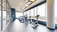 Located at 1140 Wellington St W in Ottawa's vibrant Hintonburg neighbourhood, this well-appointed fitness facility offers a thoughtfully designed space for exercise and wellness. The layout is open and functional, featuring durable grey flooring throughout and a clean, modern aesthetic defined by a neutral palette accented with bold navy blue wall details.

Floor-to-ceiling windows line the perimeter, flooding the space with abundant natural light and providing sweeping panoramic views of the Ottawa skyline and surrounding greenery — making for an inspiring workout environment. Ceiling fans and recessed panel lighting ensure consistent comfort and brightness throughout.

The facility is equipped with a comprehensive selection of cardio machines, including treadmills, upright and recumbent bikes, ellipticals, and rowing-style equipment. A dedicated strength and functional training area is separated by sleek full-height glass partition walls with aluminum framing, creating distinct zones while maintaining an open, airy feel. Structural columns with a polished concrete finish add an industrial-modern character to the space.

The entrance corridor leads seamlessly into the main workout floor through glass-panelled doors, offering a clear sightline across the entire facility. Overall, the space combines practicality with an elevated, professional atmosphere suited for a modern fitness amenity.