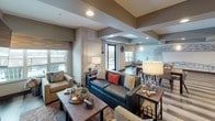 This single-floor community room is thoughtfully designed to accommodate a variety of social gatherings and everyday amenities. The space is divided into several distinct yet open zones that flow seamlessly into one another, creating a cohesive and inviting environment throughout.

The main lounge area serves as the focal point of the room, anchored by a sleek linear gas fireplace flanked by wall-mounted artwork and a large flat-screen television. Comfortable seating arrangements, including a teal leather sofa, upholstered armchairs, and wooden coffee tables, provide ample space for relaxation and conversation. Recessed lighting and coffered ceiling details add architectural interest and depth overhead.

An adjacent casual seating nook features rich hardwood-style flooring, patterned accent chairs, and a striking hand-drawn architectural mural spanning an entire wall, lending a local artistic character to the space. Natural light filters in generously through multiple windows and a glass entry door.

The fully equipped kitchen area is finished with granite countertops, tile mosaic backsplash, dark wood cabinetry, stainless steel appliances, and pendant lighting, making it well-suited for catered events or community gatherings. A pass-through window connects the kitchen visually to the broader lounge area, enhancing openness and interaction throughout the entire community room.
