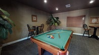 This single-floor game room is designed as a dedicated recreational space, centered around a full-size pool table with a green felt surface and rich wood frame. The room is finished with warm, taupe-toned walls and a dark ceiling, giving the space a moody, lounge-like ambiance that is reinforced by recessed overhead lighting. Patterned wall-to-wall carpeting in dark tones with a swirling motif covers the floor throughout, adding texture and visual interest underfoot.

A compact bistro-style table with two Windsor chairs occupies one corner of the room, providing a casual seating area for spectators or players between games. A wall-mounted rack for billiard accessories is positioned nearby, keeping equipment organized and accessible. Large tropical potted plants are placed throughout the room, introducing a natural element that softens the space.

French doors along one wall allow access to an outdoor area, with wood-slat blinds providing light and privacy control. Additional windows fitted with matching wood blinds further contribute to the room's warm, controlled atmosphere. A framed artwork piece decorates the wall, adding a finished, intentional touch to the overall design. The layout provides ample clearance around the pool table, making the room fully functional for gameplay.