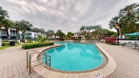 This outdoor amenity space showcases a beautifully designed resort-style swimming pool area that evokes a true Florida leisure atmosphere. The pool itself is spaciously proportioned with a smooth, curved freeform shape and striking turquoise water, reaching a depth of 6 feet. It is framed by an elegant decorative paver deck that provides ample space for lounging and socializing.

Surrounding the pool, lush tropical landscaping creates a verdant backdrop, with mature royal palm trees and vibrant flowering shrubs adding color and a sense of seclusion. Lounge chairs are arranged along the pool deck, and shaded seating areas with market umbrellas offer a respite from the sun. In the background, a well-appointed clubhouse or community building is visible, suggesting a full suite of amenities nearby.

The overall ambiance is one of relaxed, resort-inspired living — ideal for unwinding or entertaining. The combination of manicured greenery, decorative hardscaping, and carefully maintained water features creates a polished and inviting outdoor environment that stands as a signature amenity of this community.