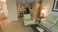 This single-floor space is designed with an open, functional layout that seamlessly connects the living and dining areas. The living area is furnished with light sage green upholstered seating, including a cushioned armchair and sofa, arranged around a sleek, dark coffee table with metal legs. A matching end table with a classic table lamp adds a warm ambient glow to the space. The flooring throughout is wall-to-wall carpeting in a neutral beige tone, contributing to a comfortable and cohesive aesthetic.

The dining area is positioned adjacent to the living space, featuring a compact dark wood table with matching chairs, ideal for intimate dining. Decorative framed artwork in coordinating tones adorns the warm beige walls, adding visual interest without overwhelming the space. A lush potted plant provides a natural accent between the two areas.

Recessed ceiling lighting illuminates the space efficiently, complementing the table lamp for layered lighting. A hallway visible from the living area leads to what appears to be a bathroom, suggesting a practical and well-organized floor plan. The overall ambiance is clean, neutral, and move-in ready.
