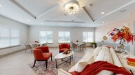 This elegantly designed single-floor community space offers a warm and inviting atmosphere through a thoughtful combination of architectural detail and curated interior finishes. The main gathering area is anchored by a striking tray ceiling adorned with a sputnik-style chandelier, lending the room a refined yet approachable character. Light herringbone-patterned hardwood flooring runs throughout, complementing the soft, neutral color palette of warm whites and subtle grays.

The open-concept layout seamlessly integrates multiple seating arrangements, including a plush sofa accented with rich terracotta throw pillows, a pair of bold rust-colored accent chairs around a round coffee table, and several dining clusters featuring tulip-style tables paired with upholstered chairs. Rows of white-shuttered windows line the walls, flooding the space with generous natural light and offering views to the outdoors.

A dedicated kitchenette area features sage-toned shaker cabinetry with glass-front upper cabinets, a striking large-format hexagonal tile backsplash, light quartz countertops, an undermount sink, and stainless steel under-counter refrigeration units. Built-in shelving provides additional display and storage space, while French doors connect to an adjacent room, enhancing the overall sense of openness and flow throughout this well-appointed gathering space.