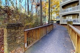 This residential community is nestled within a mature, wooded landscape that lends a distinctly natural character to the grounds. The multi-story buildings feature neutral gray siding and are fitted with private balconies on upper levels, offering elevated views of the surrounding tree canopy. The exterior architecture maintains a consistent, traditional aesthetic throughout the community.

The grounds are connected by a network of winding concrete walkways and elevated wooden boardwalks, providing pedestrian access between buildings. A notable design feature is the use of stacked natural stone — seen prominently in the retaining walls and decorative pillars that anchor the wood-railed walkway bridges. These stone elements add a rustic, crafted quality to the outdoor environment.

The landscape is dominated by tall deciduous trees, which frame the buildings and pathways with seasonal color, particularly vivid during autumn. Fallen leaves cover much of the ground, reinforcing the natural, park-like ambiance. Benches are positioned along the walkways, offering spots to enjoy the wooded surroundings. Signage is present along pathways, indicating organized community management. Overall, the setting balances built residential infrastructure with a strongly preserved natural environment.