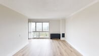 This well-appointed single-floor unit offers a clean, modern aesthetic throughout. The living area is a standout feature, characterized by light-toned hardwood flooring that extends seamlessly across the space, crisp white walls, and crown molding that adds a refined touch. A large sliding window dominates one wall, flooding the room with natural light while framing elevated views of the surrounding cityscape and greenery below. A dark-finished baseboard heating unit anchors one corner, complementing the neutral palette of the room. The open layout of the living area provides generous room for a variety of furnishing configurations, as confirmed by the 3D floor plan view. Adjacent to the living space, the kitchen is thoughtfully designed with dark espresso-toned cabinetry, white countertops, a stainless steel sink with a modern faucet, and a full-size refrigerator. The kitchen retains the same light hardwood flooring found throughout, maintaining visual continuity. A separate area near the entry features a white front door and access to additional rooms, including what appears to be a closet or secondary space. The overall ambiance is bright, neutral, and move-in ready, with a cohesive design language that balances contrast and simplicity throughout.