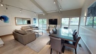 This thoughtfully designed single-floor space combines comfort and functionality in a contemporary setting. The main gathering area is anchored by dramatic vaulted ceilings with exposed beams and white tongue-and-groove paneling, complemented by industrial-style track lighting that highlights the room's generous proportions. Large sliding glass doors line the walls, flooding the interior with abundant natural light and offering views of lush greenery outside.

The lounge area features plush leather sofas arranged around a sleek marble-topped coffee table, centered on a neutral area rug over light hardwood flooring. A floor-to-ceiling stacked stone fireplace serves as a striking focal point on one wall, while a large wall-mounted television and framed artwork add visual interest throughout.

Adjacent to the lounge, a spacious dining area accommodates a full-sized glass-topped table surrounded by upholstered chairs, ideal for group gatherings. Wainscoting along the lower walls adds a refined architectural detail throughout the space.

A compact kitchenette provides practical amenities including counter space, a sink, cooktop, dishwasher, and microwave, supporting events and meetings with ease. Colorful artwork adorns the walls, adding warmth and personality to the overall aesthetic.