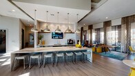Located at 5330 Sienna Pkwy in Missouri City, TX, this single-floor community clubhouse is a thoughtfully designed social hub that blends contemporary style with comfortable functionality. At the heart of the space is a striking, elongated marble-topped island bar, illuminated by a row of polished chrome pendant lights that lend an industrial-chic character to the room. The counter is lined with upholstered patterned stools, making it an ideal gathering spot. Behind the bar, a geometric hexagonal tile backsplash frames a wall-mounted TV and a built-in beverage station. The open layout transitions seamlessly into a lounge and dining area, where a rich walnut dining table, vibrant yellow accent chairs, and a bold teal area rug create an inviting atmosphere. Floor-to-ceiling windows dressed with layered patterned drapes flood the space with natural light and offer views of the outdoor patio. A dedicated media lounge features a reclaimed wood accent wall set against a white stone backdrop, anchoring multiple flat-screen TVs for an immersive viewing experience. Plank-style flooring, high ceilings, and curated décor — including sculptural planters and colorful art — complete the polished, contemporary ambiance throughout.