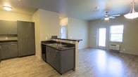 This single-floor space showcases a thoughtfully designed open-concept layout that connects the kitchen and living area in one cohesive, flowing space. The kitchen is anchored by a peninsula with dark countertops and a double stainless steel sink, complemented by sleek gray cabinetry with modern hardware. The peninsula provides both a functional workspace and a natural dividing element between the kitchen and the living area, while still maintaining an open sightline throughout. A full-size refrigerator rounds out the kitchen's appliances. The living area benefits from abundant natural light streaming in through a glass-paneled door, creating a bright and welcoming atmosphere. Light wood-look flooring extends throughout the space, adding warmth and visual continuity. A ceiling fan is installed in the living area, contributing to both comfort and airflow. The neutral wall tones throughout provide a clean, versatile backdrop well-suited for a variety of interior styles. The overall design emphasizes practicality and openness, making efficient use of the available space on a single level.