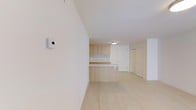 This single-floor space showcases a thoughtfully designed, minimalist interior that emphasizes clean lines and a bright, airy atmosphere. The open-concept layout seamlessly connects the living area to the kitchen, creating a sense of spaciousness and flow throughout. Light hardwood flooring runs continuously across the space, complementing the warm, natural tones of the light wood cabinetry and kitchen peninsula. The kitchen is outfitted with sleek flat-front upper cabinets and a peninsula that doubles as a casual dining or prep surface, making it both functional and visually appealing. Crisp white walls and a white ceiling reflect natural and artificial light evenly throughout, enhancing the overall sense of openness. A flush-mounted ceiling light provides subtle illumination, contributing to the space's understated elegance. A smart thermostat is conveniently mounted on the wall, pointing to modern, tech-integrated living. Entry doors with clean, simple profiles maintain the cohesive aesthetic. The overall ambiance is calm, contemporary, and well-organized, making it an inviting environment that balances practicality with modern design sensibility.