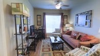 This single-floor residence offers a comfortable and thoughtfully arranged living environment. The living room serves as the heart of the space, characterized by warm hardwood flooring that adds a natural, inviting feel throughout. A ceiling fan with a built-in light fixture anchors the room from above, providing both practical air circulation and ambient lighting.

Natural light flows generously through a large central window dressed with elegant curtain panels in a rich, earthy tone, complemented by white blinds that allow for light control. The window creates a bright focal point that opens up the room visually.

The living area is furnished with a plush sofa adorned with accent pillows, a patterned armchair, and a dark wood coffee table set atop a decorative area rug with bold geometric patterns. A media console along one wall houses a flat-screen television and is accompanied by bar-height seating, adding a versatile entertaining element to the space.

Wall art and framed prints are thoughtfully placed to add character and depth to the neutral-toned walls. An open shelving unit near the entryway provides additional storage and display space. The overall ambiance is warm, casual, and welcoming, making this a functional and aesthetically cohesive living environment.