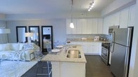This single-floor space showcases a thoughtfully designed open-concept layout that integrates the kitchen, dining, and living areas into one cohesive environment. The kitchen is a standout feature, equipped with crisp white cabinetry that extends to the upper walls, providing generous storage. Speckled granite countertops line both the perimeter workspace and a central island, which doubles as a breakfast bar with seating for acrylic ghost chairs, adding a contemporary touch. A stainless steel undermount sink sits within the island, complemented by a polished faucet. Stainless steel appliances, including a gas range, complete the culinary space. Overhead, a combination of pendant lighting above the island and track lighting ensures the kitchen is well-illuminated. Dark hardwood flooring runs throughout, grounding the light-toned cabinetry and counters. The open flow into the living and dining areas creates a spacious, airy ambiance, with the neutral gray walls contributing to a clean, modern aesthetic. A large floor mirror in the living area enhances the sense of depth and light throughout the space.