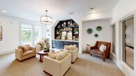 Located at 1540 Edgefield Dr in Garland, Texas, this single-floor living room is a well-appointed space that balances comfort and functionality with refined interior design. The room's focal point is a striking dark-stained built-in shelving unit framed by an arched architectural detail, complete with lower cabinetry for storage and open upper shelves displaying books, greenery, and decorative accents. A drum pendant light fixture anchors the main seating area, which features a curated mix of plush cream upholstered chairs, a warm-toned leather armchair, and a round wood coffee table — creating an inviting conversation arrangement. Neutral wall-to-wall carpeting and crisp white walls provide a clean, versatile backdrop, while decorative green resin wall art adds a distinctive pop of color and texture. A built-in desk nook with dark cabinetry, a task light, and a sage green accent wall offers a dedicated workspace tucked seamlessly into the layout. Large windows along one wall allow natural light to flow through the room, offering views of the surrounding greenery outside. The overall ambiance is warm, polished, and welcoming, making this a highly functional and aesthetically cohesive living space.