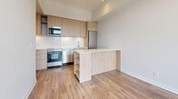 This thoughtfully designed one-bedroom, one-bathroom unit makes efficient use of its compact footprint across a single floor. Light hardwood flooring runs throughout, creating a cohesive and warm aesthetic that ties each space together seamlessly.

The open-concept kitchen is a standout feature, outfitted with light wood cabinetry, a white countertop, and a full suite of stainless steel appliances including a built-in microwave, dishwasher, refrigerator, and range. A peninsula with open shelving below extends into the living area, providing additional prep and storage space while maintaining an open feel.

The living area is bathed in natural light courtesy of expansive floor-to-ceiling windows that frame striking panoramic views of the city skyline — a defining characteristic of this elevated unit. The bedroom continues this theme with its own large window wall offering equally impressive urban vistas.

The hallway connects the living spaces to the bedroom and bathroom, and features a built-in shelving unit for additional storage. The full bathroom is finished with clean white subway tile surrounding a soaker tub and shower combination, complemented by chrome fixtures and tile flooring. The overall design palette — white walls, natural wood tones, and modern finishes — gives the unit a bright, contemporary, and move-in-ready appeal.