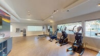 This fitness center is designed to support a comprehensive workout experience within a single, open-plan floor layout. The space is finished with light hardwood-style flooring throughout, contributing to a clean and modern aesthetic that is both functional and inviting.

The cardio section is prominently featured and includes treadmills with individual screens, an elliptical trainer, a stationary cycle, and a stair climber. These machines are positioned near a continuous row of wide windows, allowing natural light to fill the space and providing outdoor views during workouts. A dedicated treadmill alcove with surrounding windows offers an especially bright workout environment.

The strength training area includes a multi-function cable machine with an attached workout guide, a flat weight bench, and a rack of dumbbells in varying sizes. A water station is conveniently mounted nearby. A large wall mirror anchors one side of the room, visually expanding the space and supporting form-checking during exercise.

Additional fitness accessories, including exercise balls and resistance tools, are stored on a dedicated rack. Track lighting runs along the ceiling, providing even illumination throughout. The fitness center also includes a dedicated entry door, posted fitness center guidelines, and a hand sanitizer station, reflecting attention to user safety and facility management.
