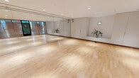 Located on Tresser Boulevard in Stamford, Connecticut, this fitness and movement studio offers a generously sized, open-plan space designed to accommodate a wide range of exercise and wellness activities. The studio is defined by its polished hardwood flooring, which runs the full length of the room and provides an ideal surface for yoga, dance, fitness classes, and more.

A standout feature of the space is its striking ombre accent wall, transitioning seamlessly from warm terracotta tones at the top to cool ocean blues at the base, adding a vibrant and calming visual element to the environment. Opposite this wall, floor-to-ceiling mirrors extend across the full width of the room, visually expanding the space and providing essential functional reflection for movement-based activities.

Recessed lighting throughout the ceiling ensures bright, even illumination across the entire studio. The entry side of the room is lined with elegant sheer curtains that soften the transition from the exterior glass partition, allowing natural light to filter in while maintaining a sense of privacy. Built-in white cabinetry along one wall provides practical storage, and subtle greenery adds a refreshing, natural touch. The overall ambiance is clean, modern, and welcoming — purpose-built for focused movement and wellness practice.